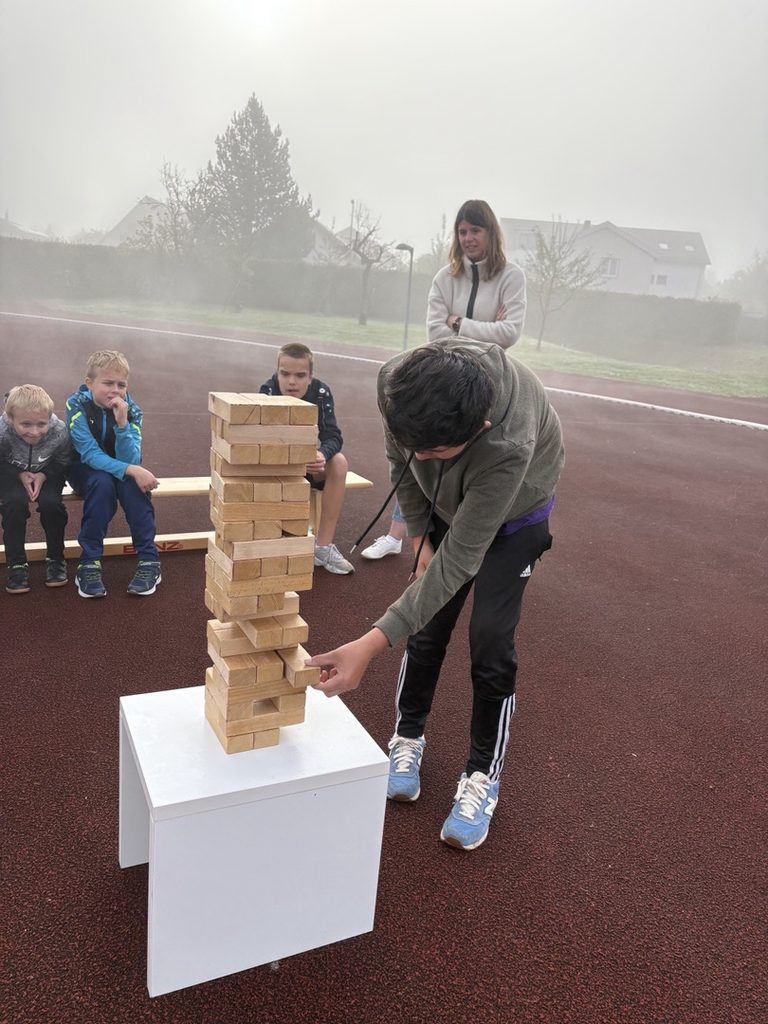 Ein anderer Schüler zieht einen Stein aus einem Jenga- Turm, andere schauen zu und fiebern mit