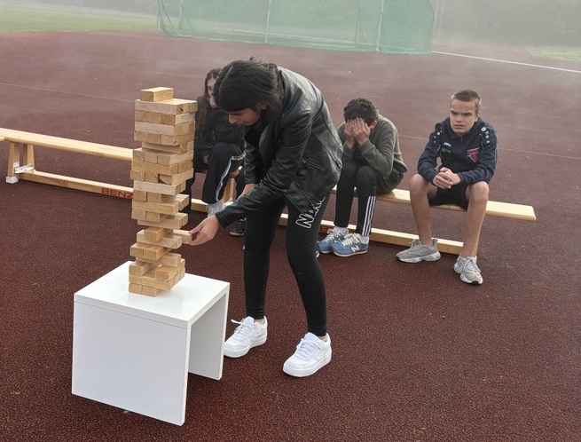 Eine Schülerin zieht einen Stein aus einem Jenga- Turm, andere schauen zu und fiebern mit